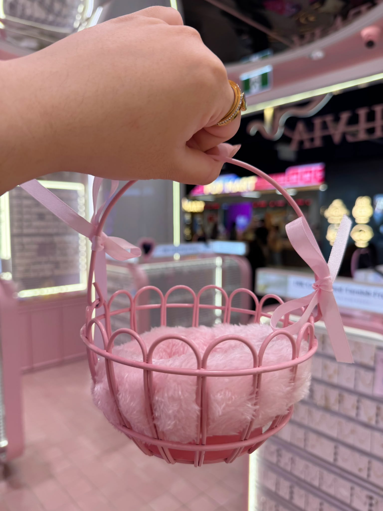 A hand holding a small pink metal basket with a fluffy pink lining and ribbon handles, photographed in a brightly lit retail store interior.